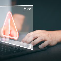 Laptop billentyűzetén gépelő kezek piros figyelmeztető jellel és technológiai ikonokkal.